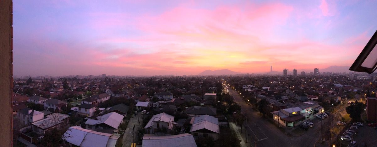 A panoramic view captures a vibrant sunset over a bustling urban landscape. The sky is filled with hues of pink and orange, transitioning to softer shades as the horizon stretches. The silhouette of distant mountains and city buildings adds depth to the scene.