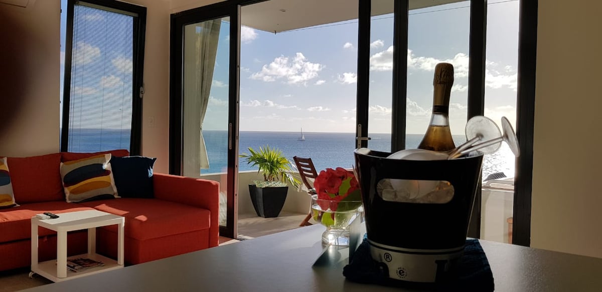 The living area features a red sectional sofa, a coffee table, and large glass doors that overlook the ocean. A bottle of champagne is placed in an ice bucket, accompanied by flowers on the table. The view includes the sea and distant sailboats, enhanced by natural light.
