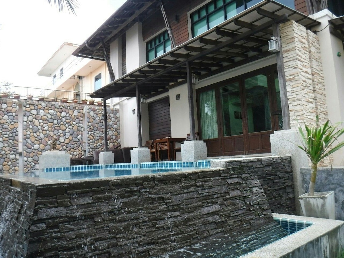 The image shows a modern architectural structure featuring a stone wall with a cascading water feature. A spacious covered patio is visible, with wooden furniture partially seen through large glass doors. Lush greenery and a decorative plant accentuate the outdoor area.