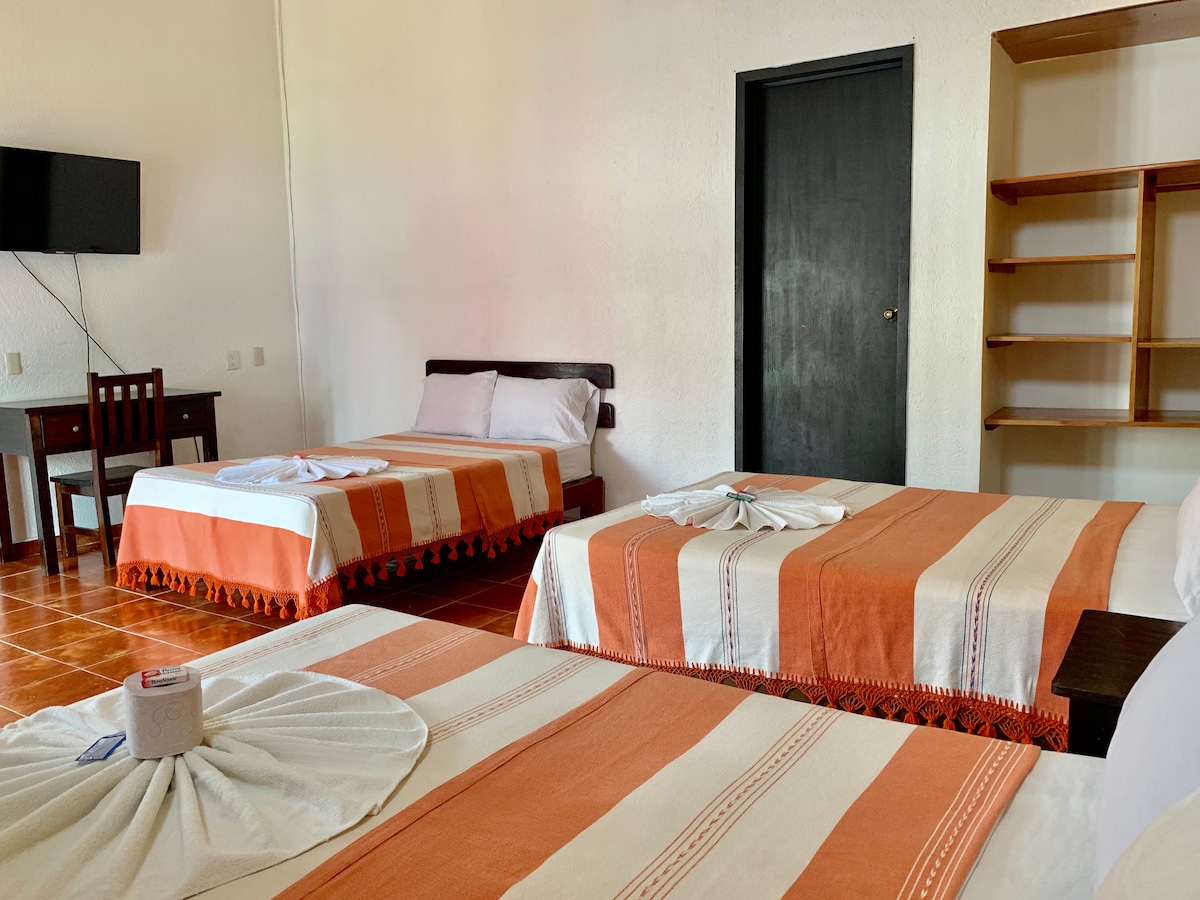 The room features three spacious beds, each adorned with striped orange and white blankets. A wall-mounted television is visible, along with a simple wooden shelving unit. Natural light from a nearby window enhances the light-colored walls and tiled flooring.