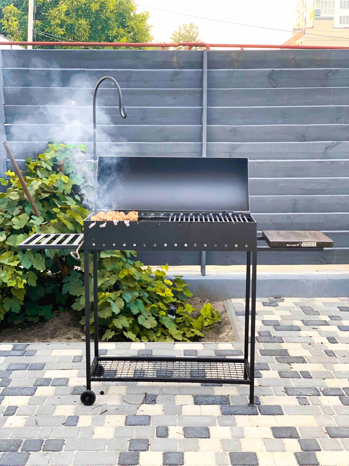 A black charcoal grill sits in a private courtyard with smoke gently rising. The grill features a cooking surface and a side shelf for food preparation. Lush greenery can be seen in the background, complementing the paved area surrounding the grill.