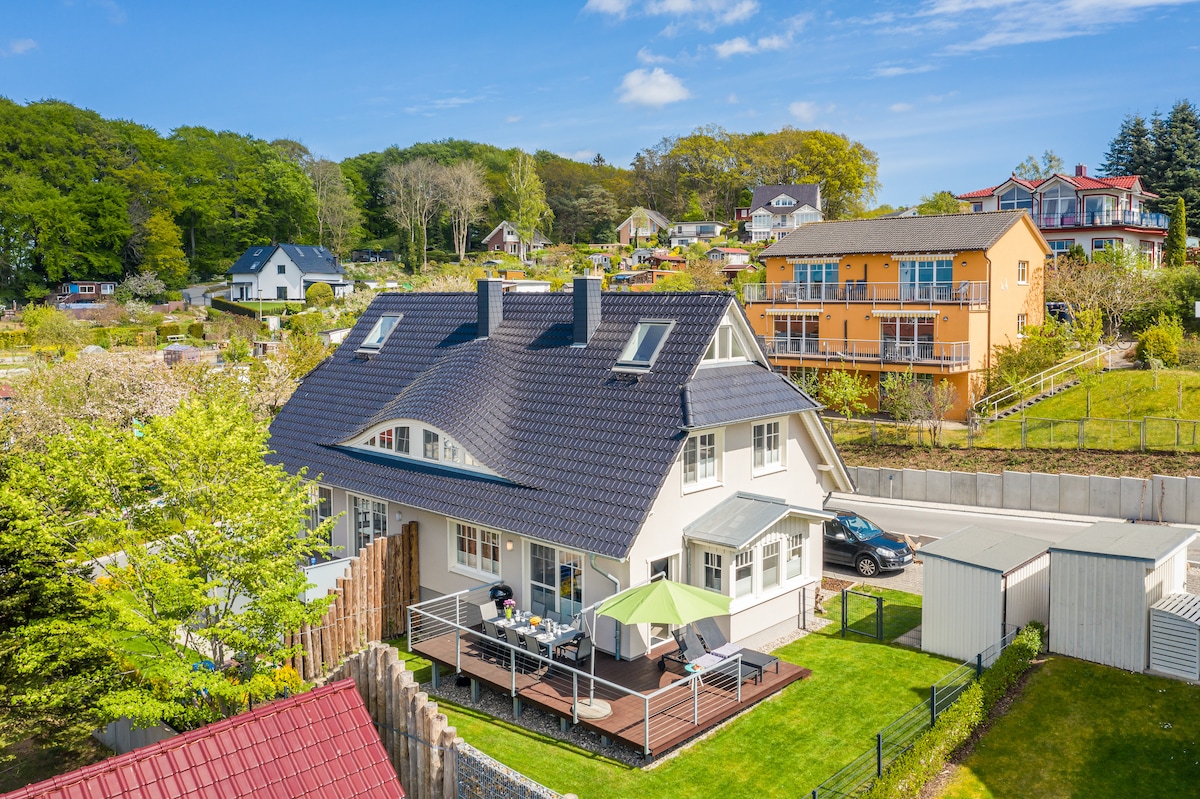 An aerial view of a spacious holiday house surrounded by greenery. A large deck with an umbrella is visible in the front yard, complemented by well-maintained gardens. The surrounding area features other residential buildings nestled among trees, creating a serene atmosphere.