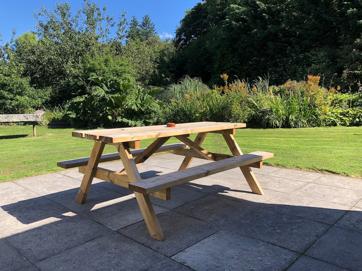 A wooden picnic table is positioned on a paved patio, surrounded by well-maintained grass. Lush greenery and flowering plants can be seen in the background, providing a natural and serene outdoor setting.