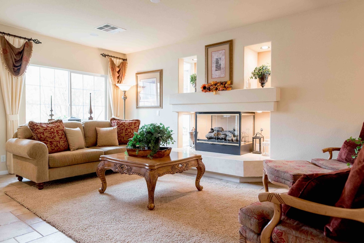 The living room features a pair of plush sofas, one adorned with decorative cushions, positioned near a modern fireplace. A wooden coffee table sits centrally on a soft area rug, complemented by potted plants and tasteful wall décor. Natural light streams through a large window.