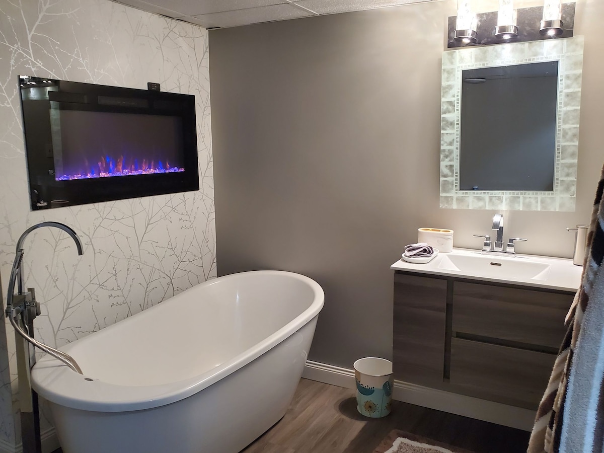 A modern bathroom features a freestanding soaking tub and a sleek vanity with a contemporary sink. A wall-mounted fireplace provides gentle illumination, enhancing the space's tranquil ambiance. A large mirror reflects the room's design, while subtle tones of gray create a calming atmosphere.