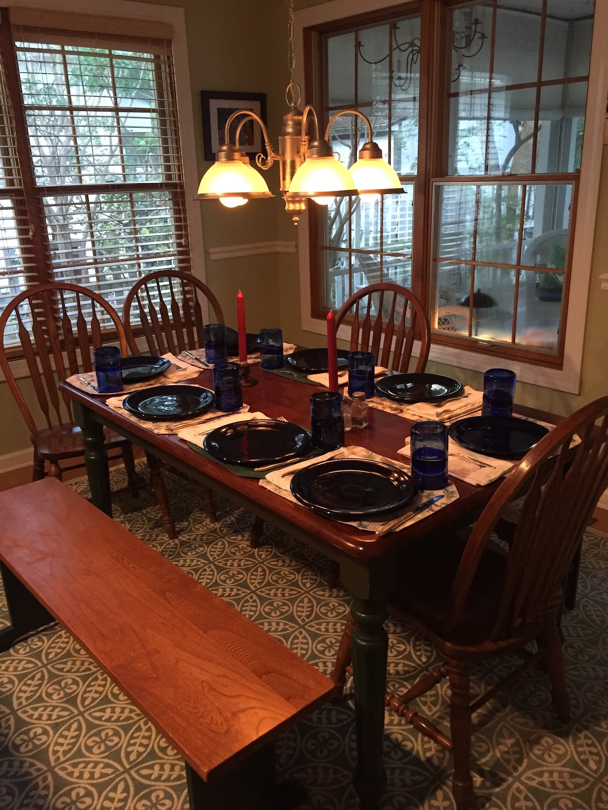 A dining table is set with eight place settings, featuring dark plates and blue glasses. Two red candles are placed in the center, enhancing the warm ambiance. The space is brightened by natural light from nearby windows, with a patterned rug underneath.