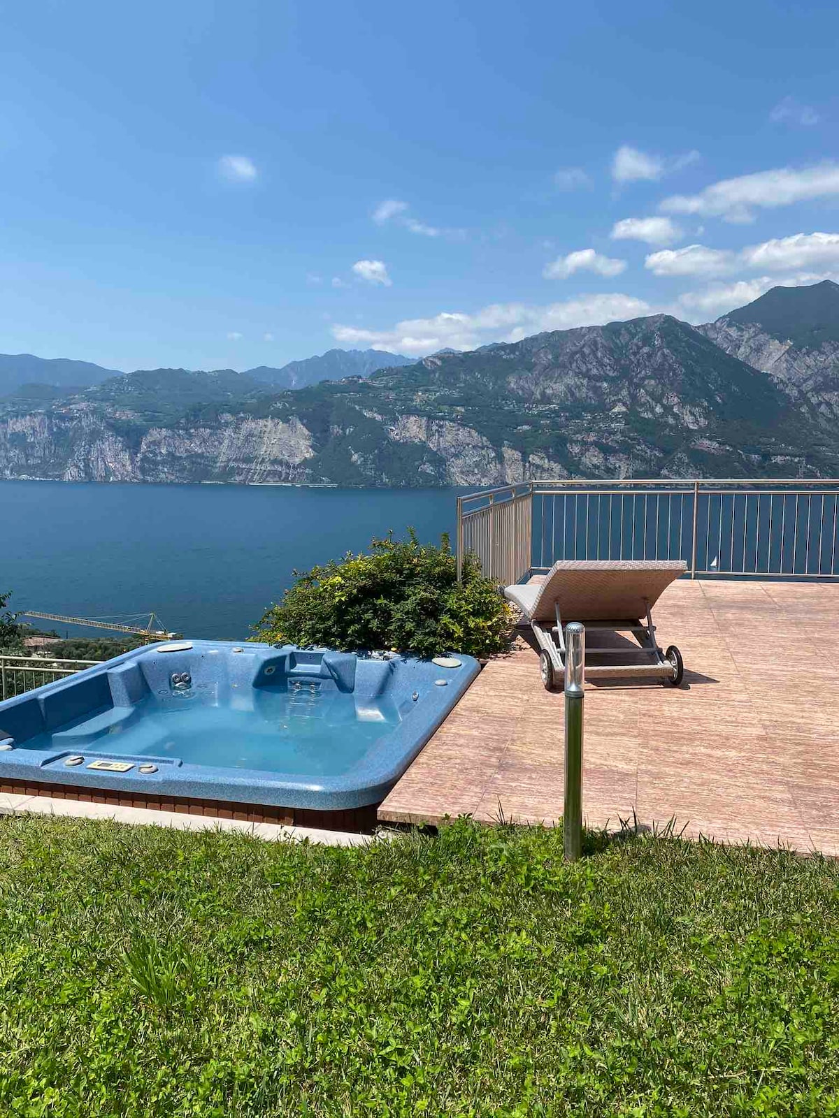 A private hot tub is positioned on a patio overlooking Lake Garda, surrounded by lush greenery. A sun lounger is placed nearby, creating a space for relaxation under the clear blue sky. The stunning mountain backdrop enhances the tranquil environment.