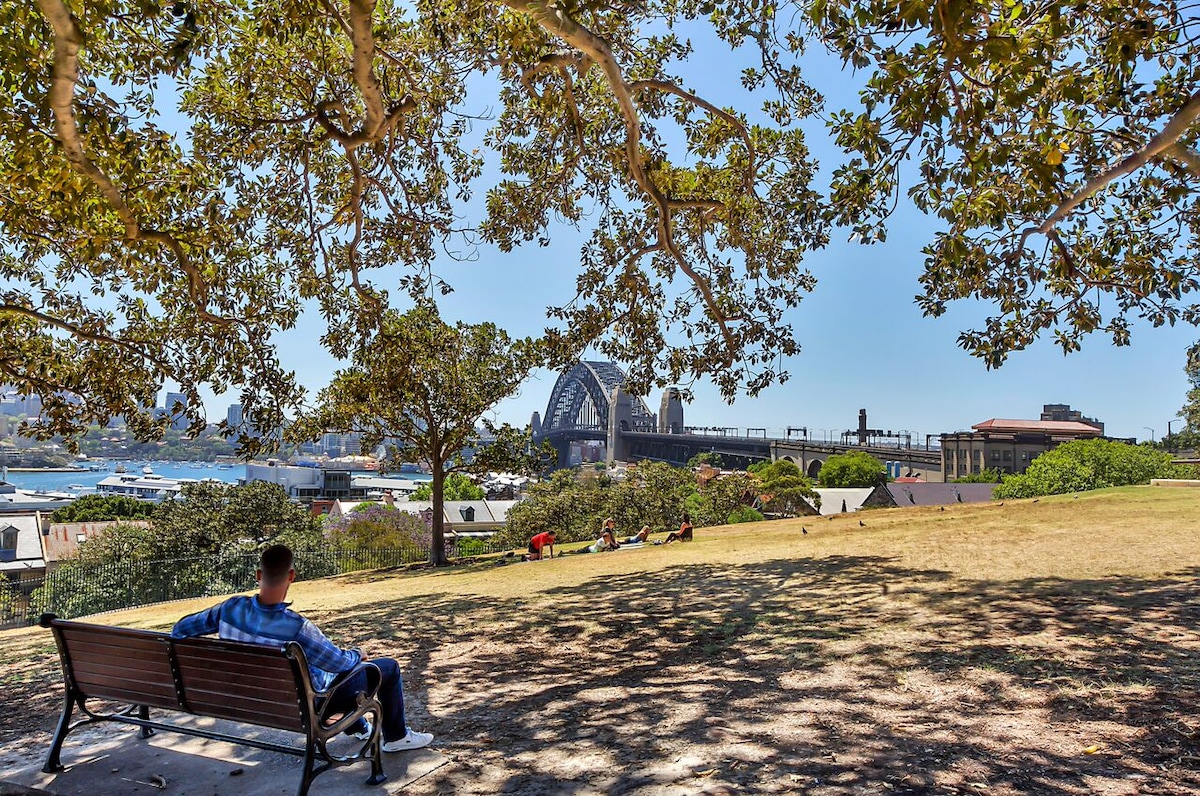 A peaceful park setting is depicted, featuring a man seated on a wooden bench under the shade of large trees. The view includes the Sydney Harbour Bridge in the background, with water and a few people enjoying the grassy area.