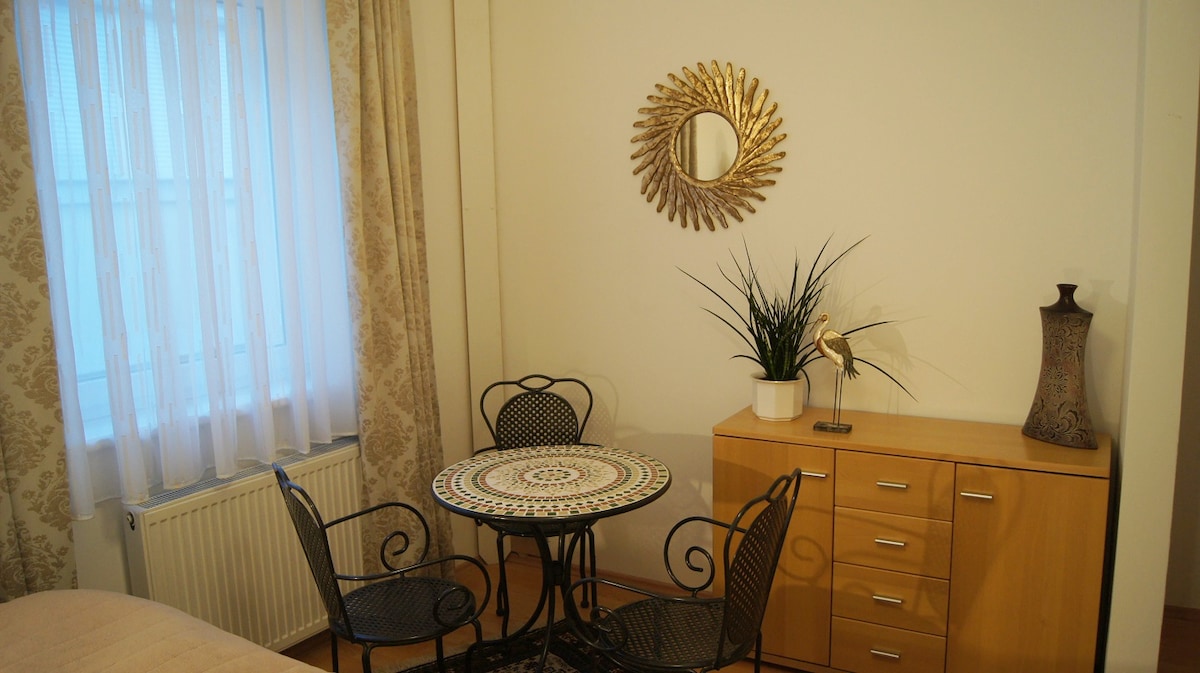 A small dining area with a decorative round table is surrounded by four black chairs. A glass vase and a potted plant are displayed on a wooden cabinet, while a round mirror adorns the wall. Sheer curtains allow soft light to enter the room.