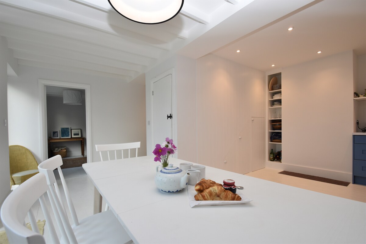 A light-filled dining area features a white table set with pastries and a small floral arrangement. Soft, neutral-colored walls complement the wooden beams above. A doorway leads to a cozy corner with a chair, while a blue kitchen area is subtly indicated in the background.