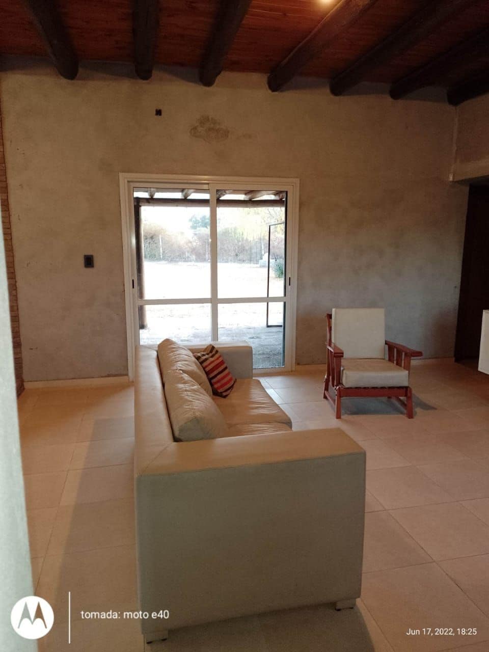 A light-filled living area is presented, featuring a comfortable sofa and a wooden armchair. Large glass doors open to the outdoor space, allowing for natural light to brighten the neutral-colored walls and tiled floor.