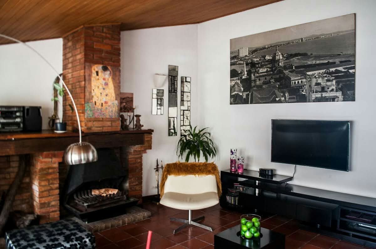 An inviting living room features a classic brick fireplace and a large black-and-white photograph on the wall. A cozy white chair sits near the fireplace, complemented by a sleek black media console and a colorful bowl of green fruits on a coffee table.