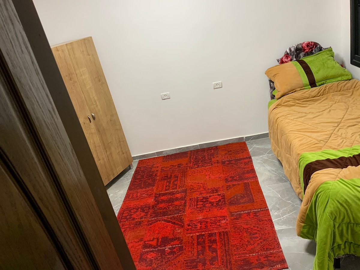 A simple bedroom features a spacious bed adorned with a green and brown blanket. A vibrant red rug lies on the floor, adding a touch of color. A modest wooden wardrobe stands in the corner, and natural light floods the space through a window.