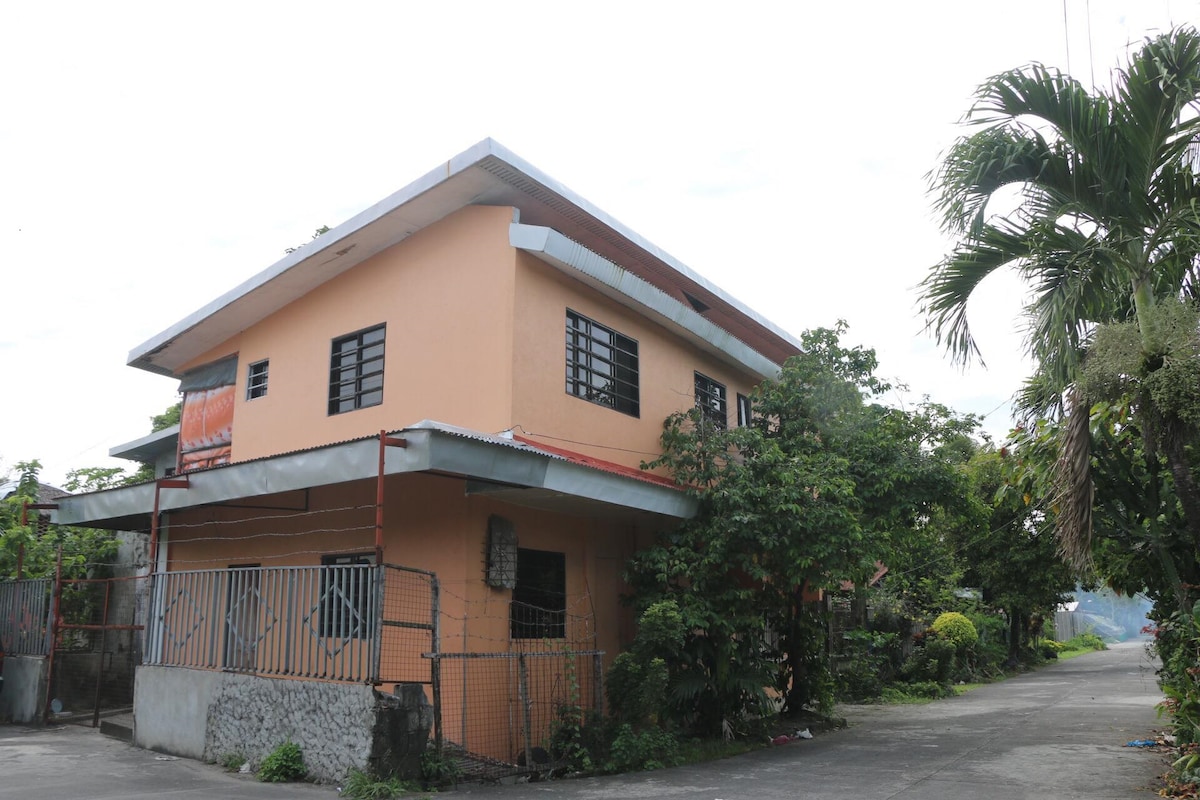 The exterior of the two-story apartment features an orange facade surrounded by greenery. Windows with metal bars provide ample light, while the covered porch adds functionality. A pathway leads to the building, flanked by lush plants and trees, contributing to a tranquil atmosphere.