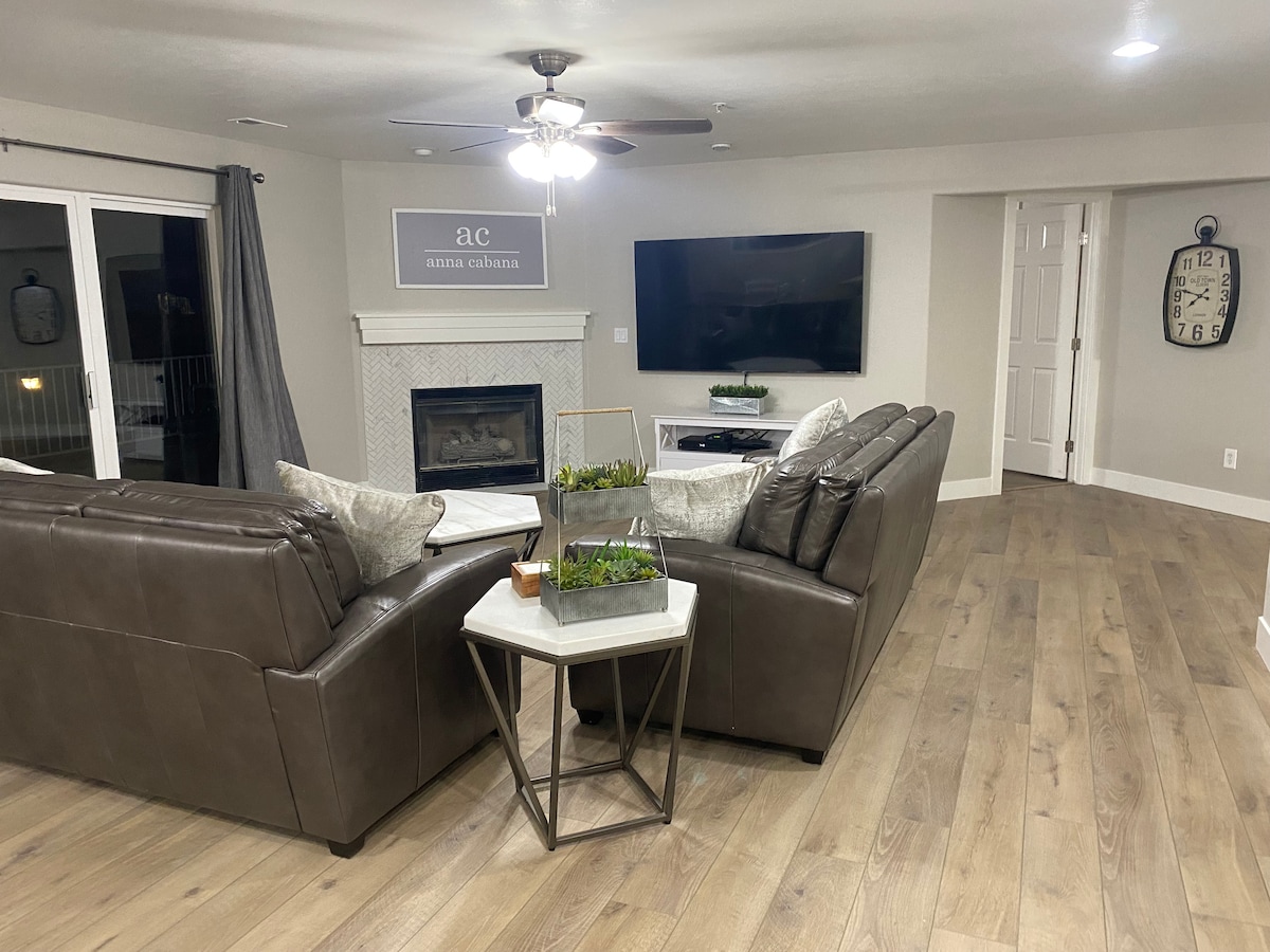 A spacious living area features two leather sofas arranged around a coffee table, with a modern gas fireplace as a focal point. A large flat-screen TV is mounted on the wall opposite, while natural light pours in through glass doors leading to a balcony.