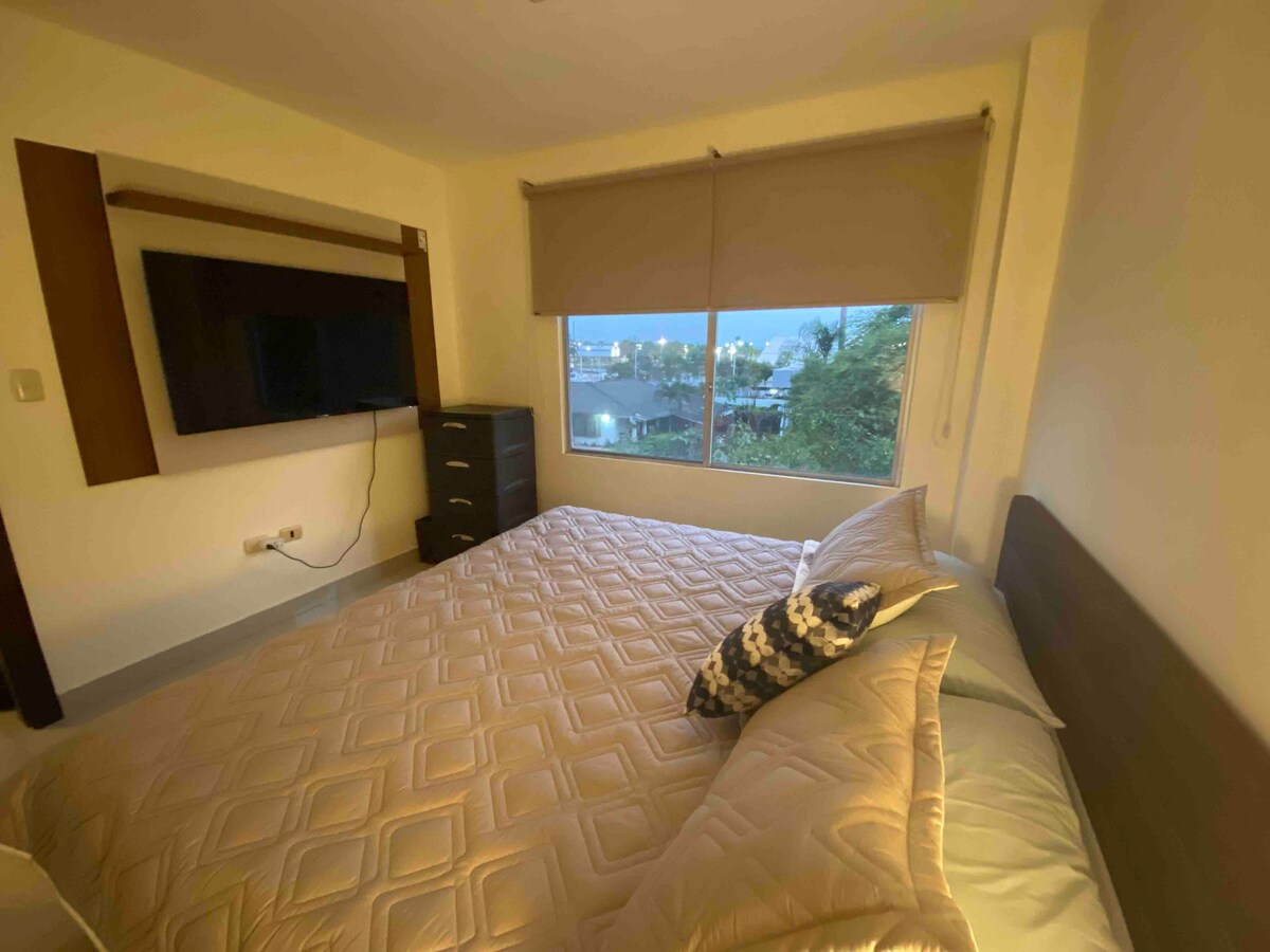 A spacious bed is positioned next to a large window, offering views of the surroundings. A television is mounted on the wall across from the bed. A dark wooden dresser provides additional storage, and soft natural light fills the serene space.
