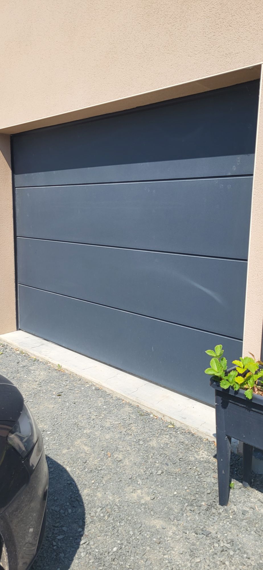 A sleek, modern garage door with horizontal panels is shown. The door is positioned against a light-colored wall, with a gravel surface visible in the foreground. A planter with greenery is also present beside the door.