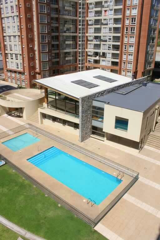 An aerial view showcases a well-maintained outdoor area featuring two swimming pools surrounded by a paved deck. Adjacent is a modern building with large windows and a stone facade, set against a backdrop of lush green grass.