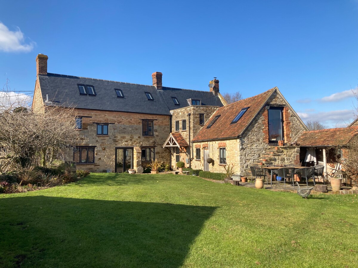 A spacious stone house is framed by a well-maintained garden, featuring manicured lawns and various outdoor seating arrangements. The building displays a mix of traditional and modern architectural elements, with large windows reflecting sunny skies and inviting views of the countryside.