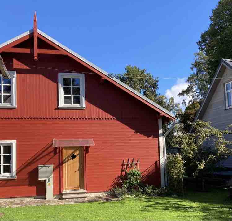 The exterior of a red wooden house is visible, featuring a pitched roof and white-framed windows. A wooden door is positioned centrally, with a small roof overhang. The surrounding area includes green grass and a couple of shrubs, under a clear blue sky.
