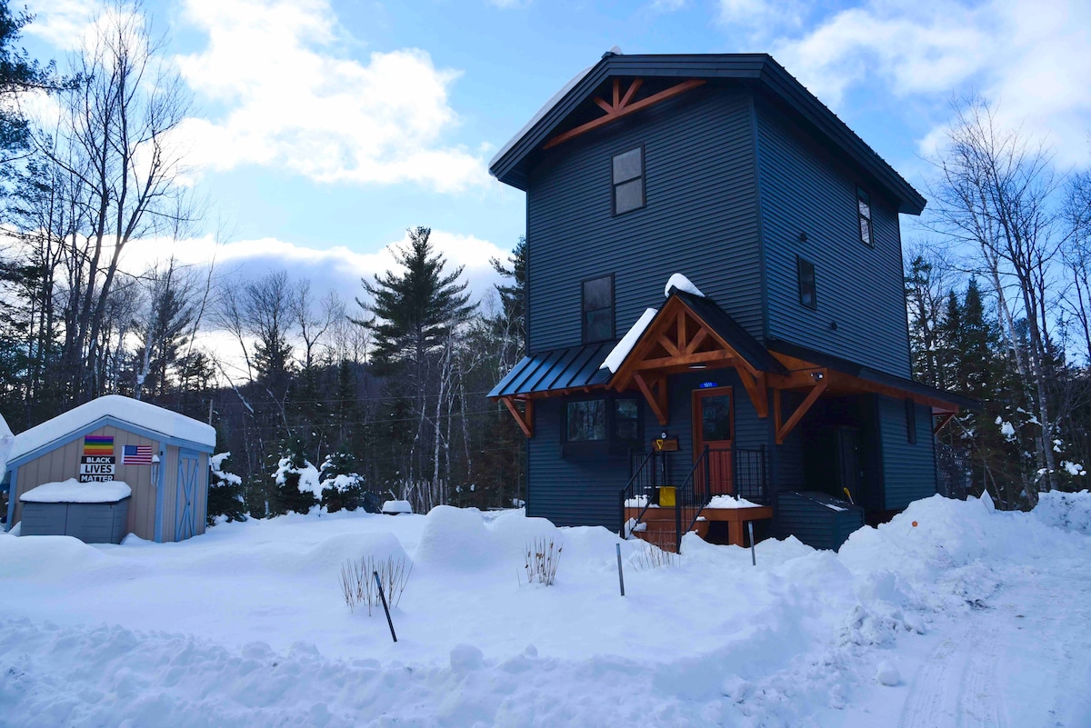 A modern three-story home features a striking dark exterior complemented by wooden accents. Its inviting entrance is framed by an overhang, while snow blankets the surrounding landscape. Adjacent, a shed is visible, adding to the property’s functionality.
