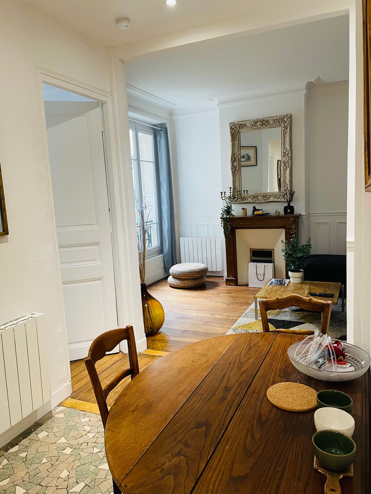 A dining area is visible, featuring a wooden table and curved chairs. A mirror hangs on the wall opposite a decorative fireplace. Natural light enters through a window, illuminating the wooden floor and a cozy seating arrangement in the adjacent room.