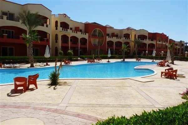 The image depicts a spacious swimming pool curving through a landscaped area, surrounded by palm trees and lounge chairs. The pool's clear blue water reflects the sunny sky, while the colorful building in the background adds vibrancy to the scene.
