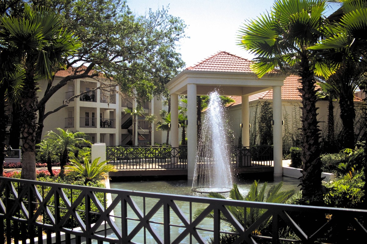 A scenic outdoor view showcases a tranquil fountain gently spraying water into a reflecting pool. Lush green palm trees and vibrant landscaping surround the area, complemented by a tastefully designed covered structure with a red-tiled roof in the background.