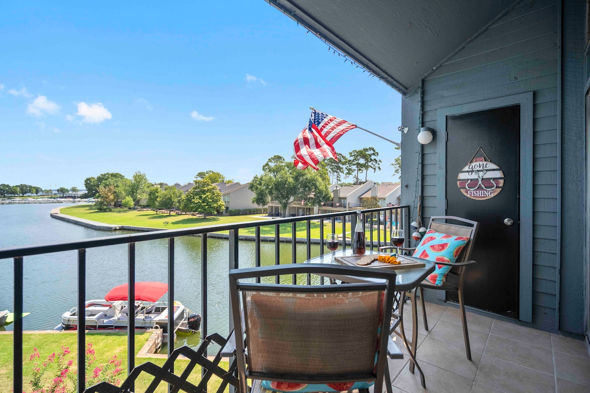 A private balcony overlooks the marina, featuring a small dining table set with colorful dishes. An American flag gently flutters in the breeze, while lush greenery and boats are visible along the water's edge.