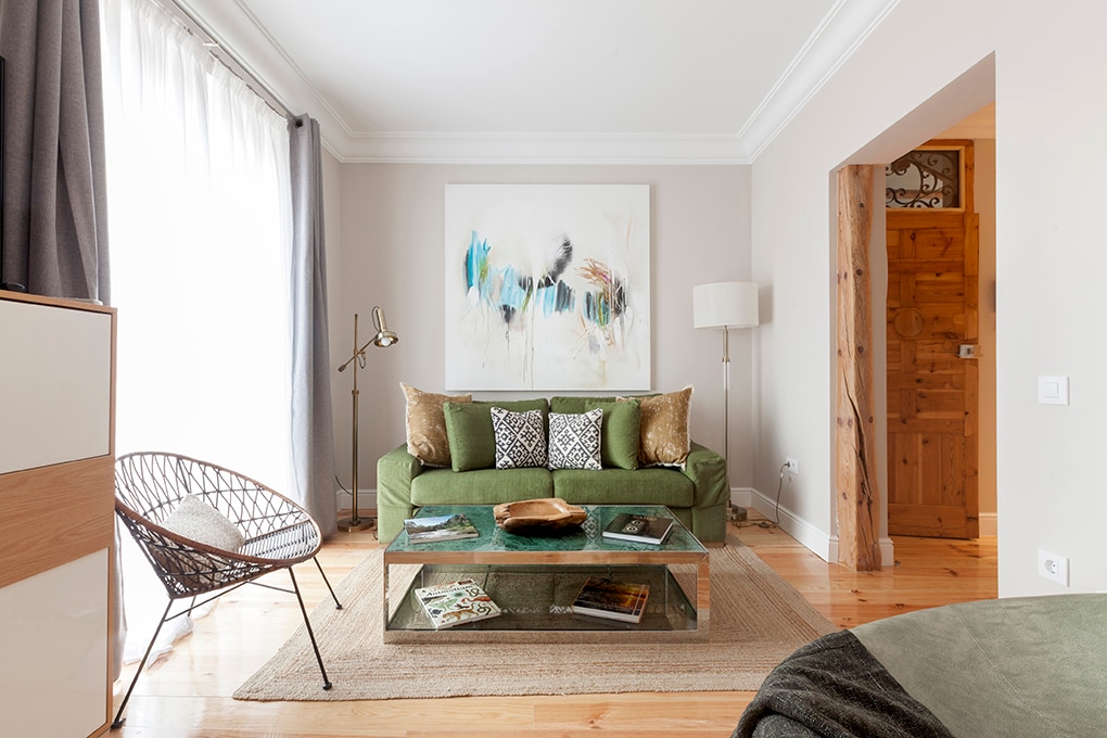 A bright living room is showcased, featuring a green sofa adorned with decorative cushions. A glass coffee table rests on a neutral rug, accompanied by an art piece above. Sunlight filters through sheer curtains, illuminating wooden floors and highlighting the warm ambiance.