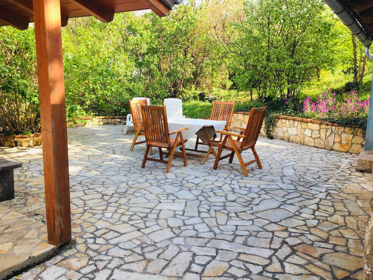 An outdoor seating area is featured on a stone-paved patio, surrounded by lush greenery. A wooden table is accompanied by four chairs, creating an inviting space for dining or relaxation. Colorful flowers can be seen in the nearby garden, enhancing the natural surroundings.
