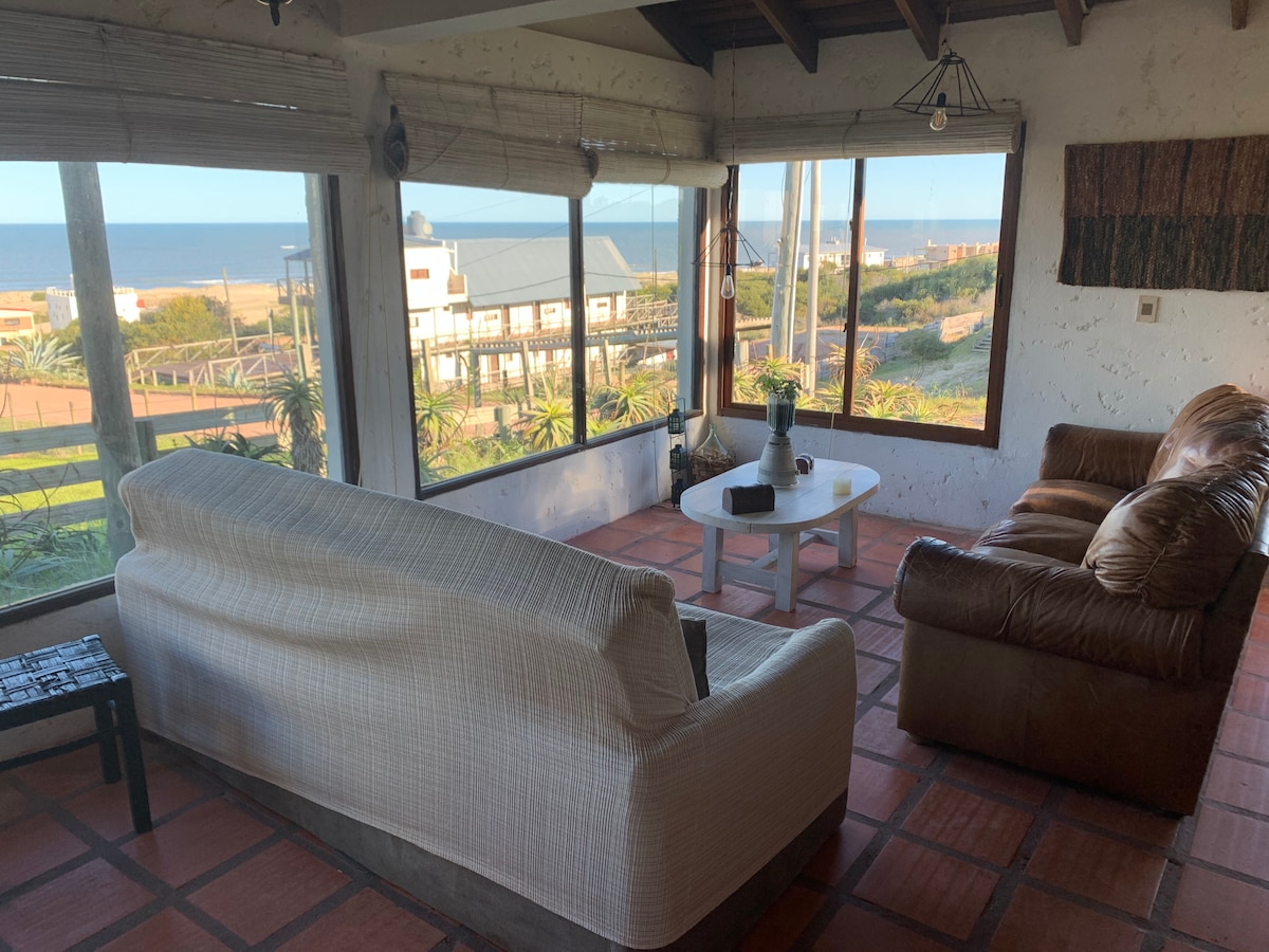 A sunlit living area with large windows offers a view of the ocean. Furnishings include a white sofa and a brown leather couch, complemented by a round coffee table. A simple vase with greenery sits on the table, enhancing the inviting space.