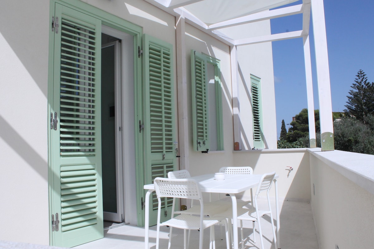 A terrace features a white table and four chairs, surrounded by light green shutters on the adjacent doors. The space is well-lit by natural sunlight, with a clear blue sky visible above. The white railing adds a clean, modern touch.