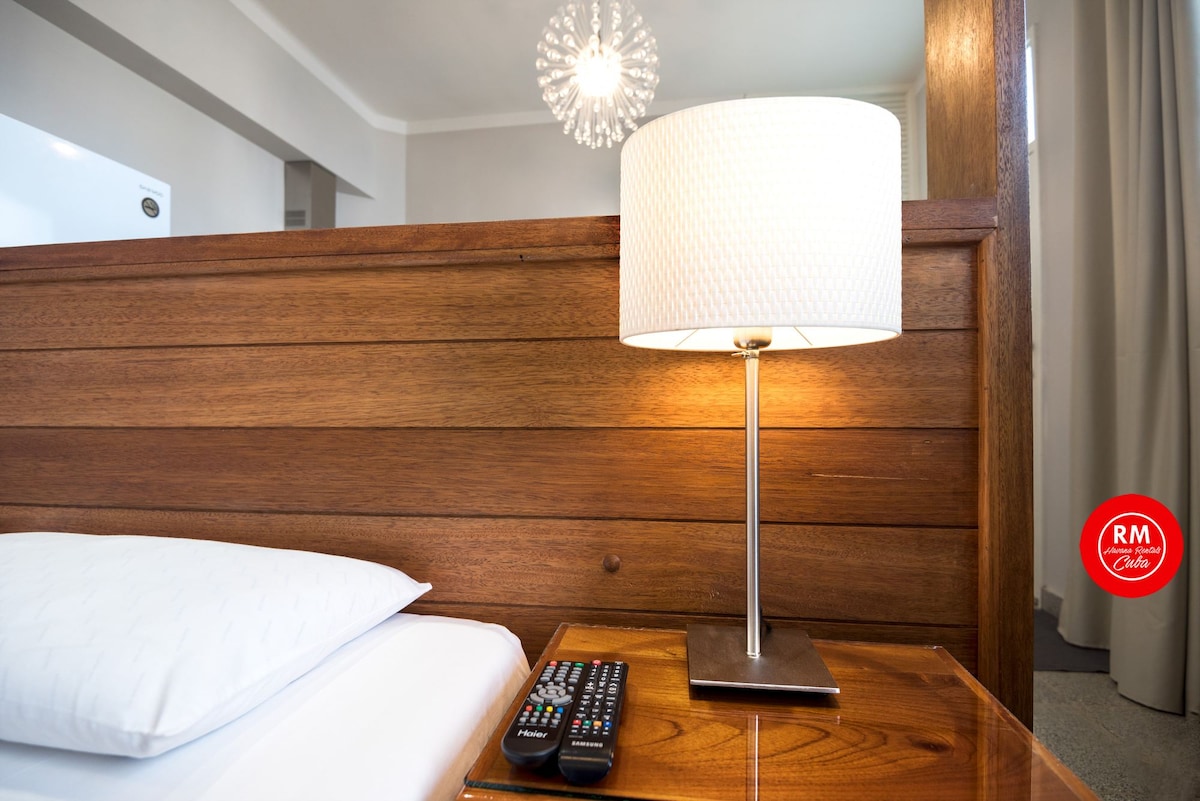 A bedside table is shown with a modern lamp and a remote control resting on its surface. A wooden headboard adds a touch of warmth to the cozy sleeping area, with soft bedding partially visible in the frame.