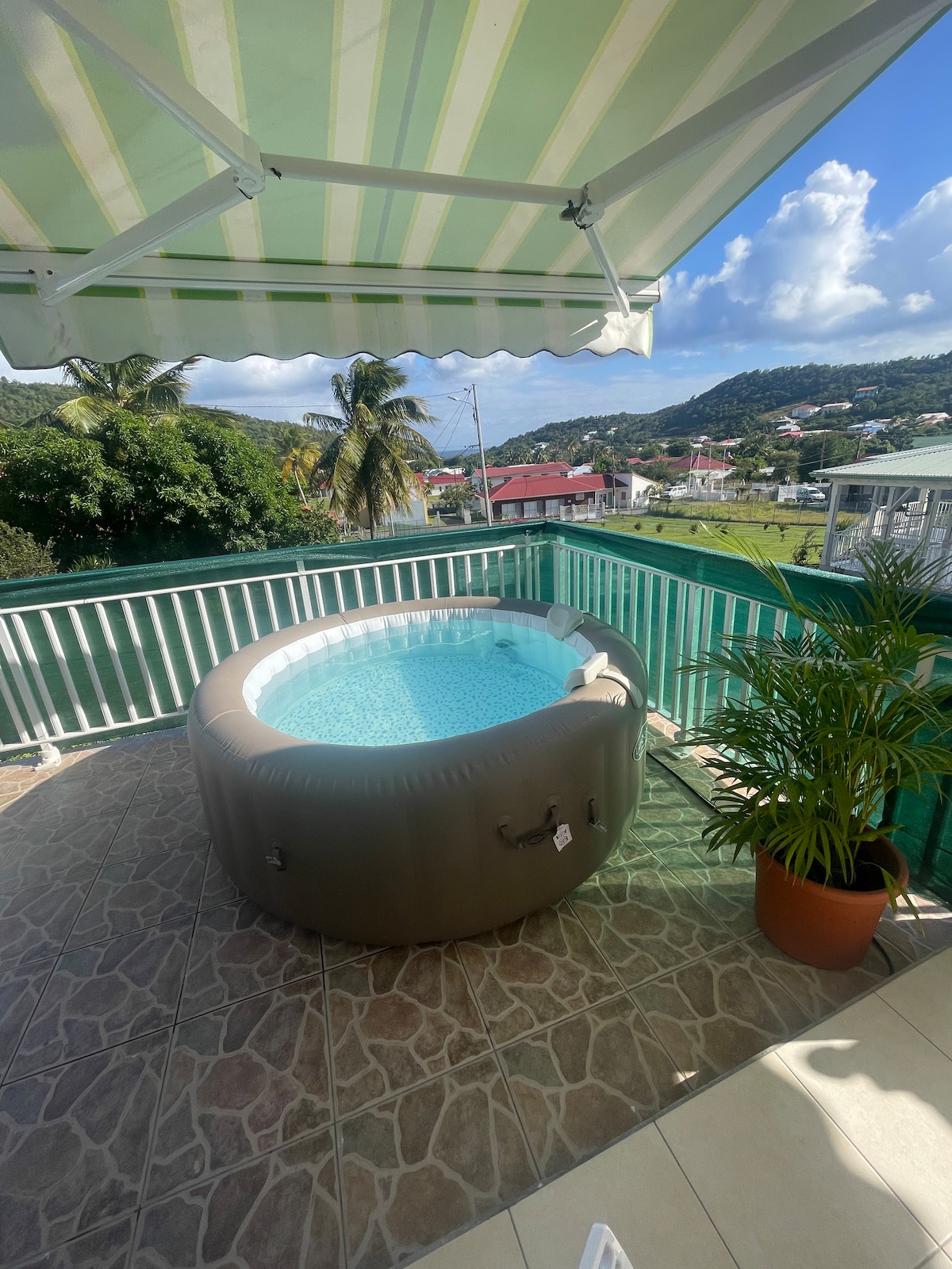 A round jacuzzi is positioned on a terrace, surrounded by tiled flooring. Lush greenery and distant hills are visible beyond a railing. The canopy above provides shade, while sunlight reflects off the clear water, creating a serene setting.