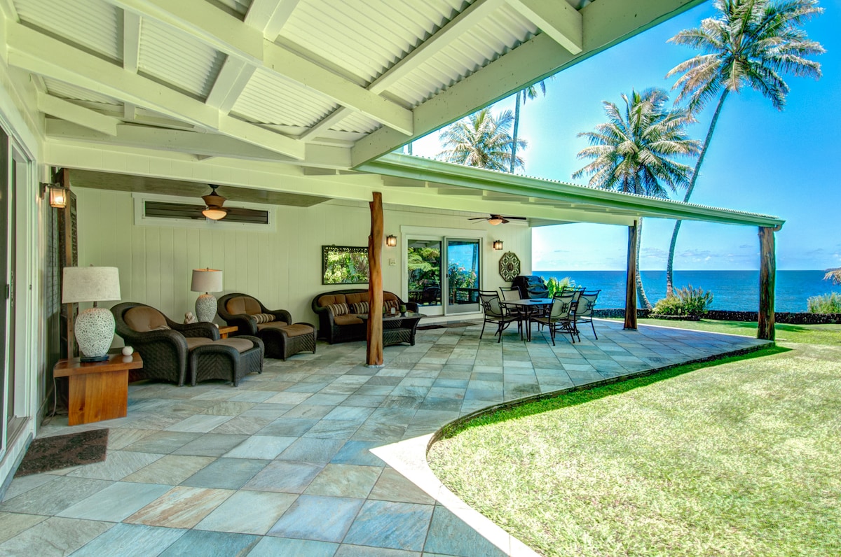An open patio features a smooth stone floor, comfortable seating with wicker chairs, and a dining table. Lush green grass surrounds the area, leading to a view of palm trees and the ocean in the background beneath a clear blue sky.