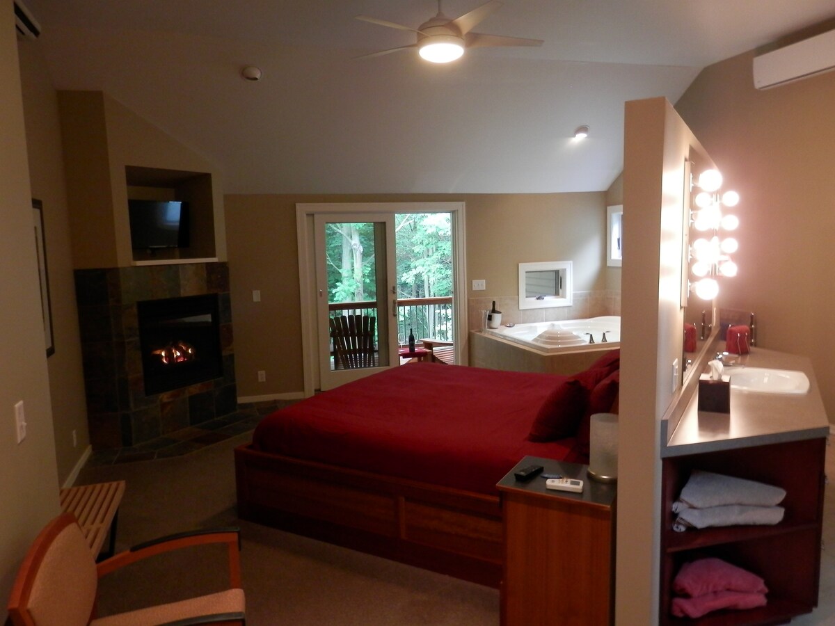 The suite features a spacious bed covered in deep red bedding, complemented by a stylish wooden frame. A cozy seating area is visible beside a gas fireplace. A large window provides views of the forested surroundings, while a whirlpool tub and a well-lit vanity are situated nearby.