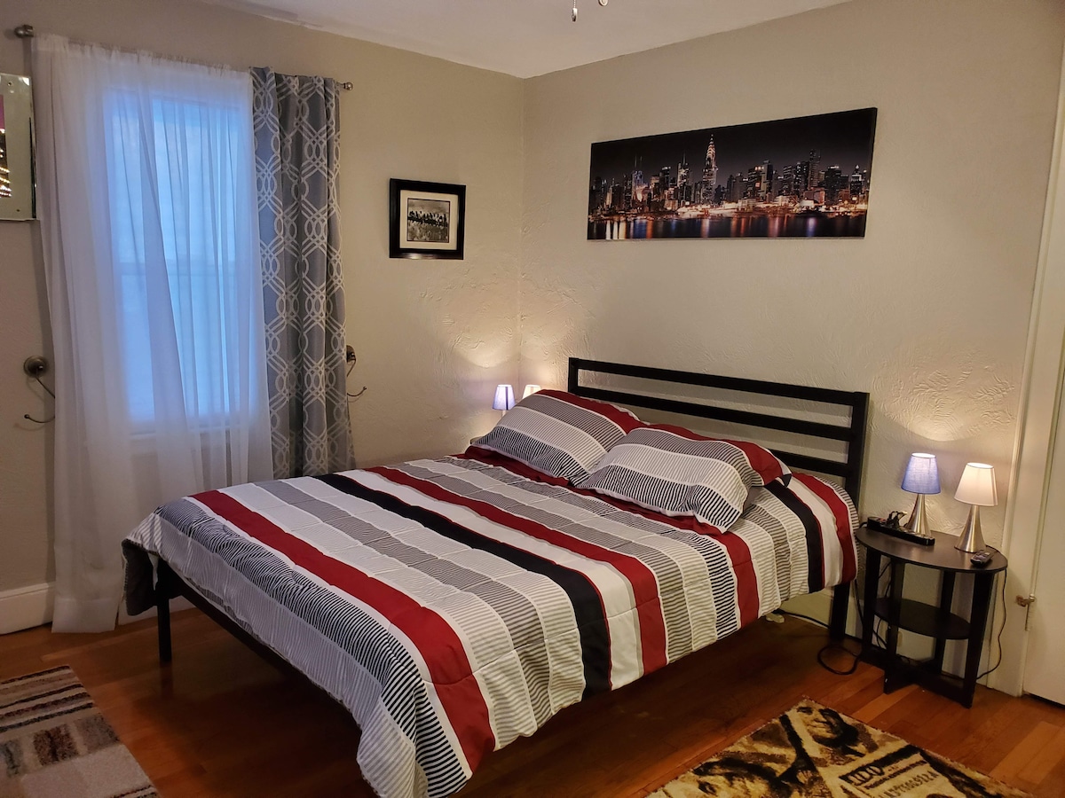 A queen bed is centered in the room, adorned with striped bedding in shades of red and gray. Two small bedside lamps provide soft lighting, and a window is draped with sheer white curtains. A scenic New York skyline print hangs above the bed.