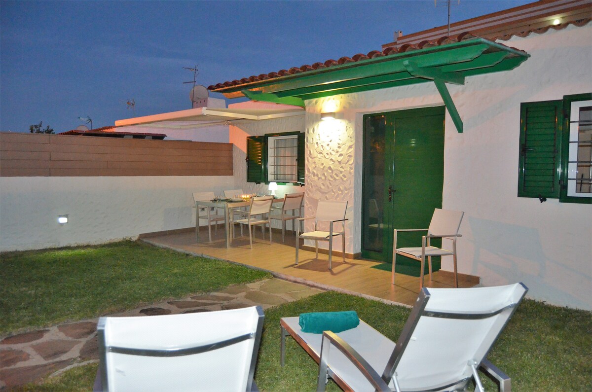 An outdoor seating area features several chairs arranged around a small table, set against a backdrop of a white bungalow. A green awning provides shade, while a well-maintained lawn surrounds the space, offering a relaxed and welcoming environment.