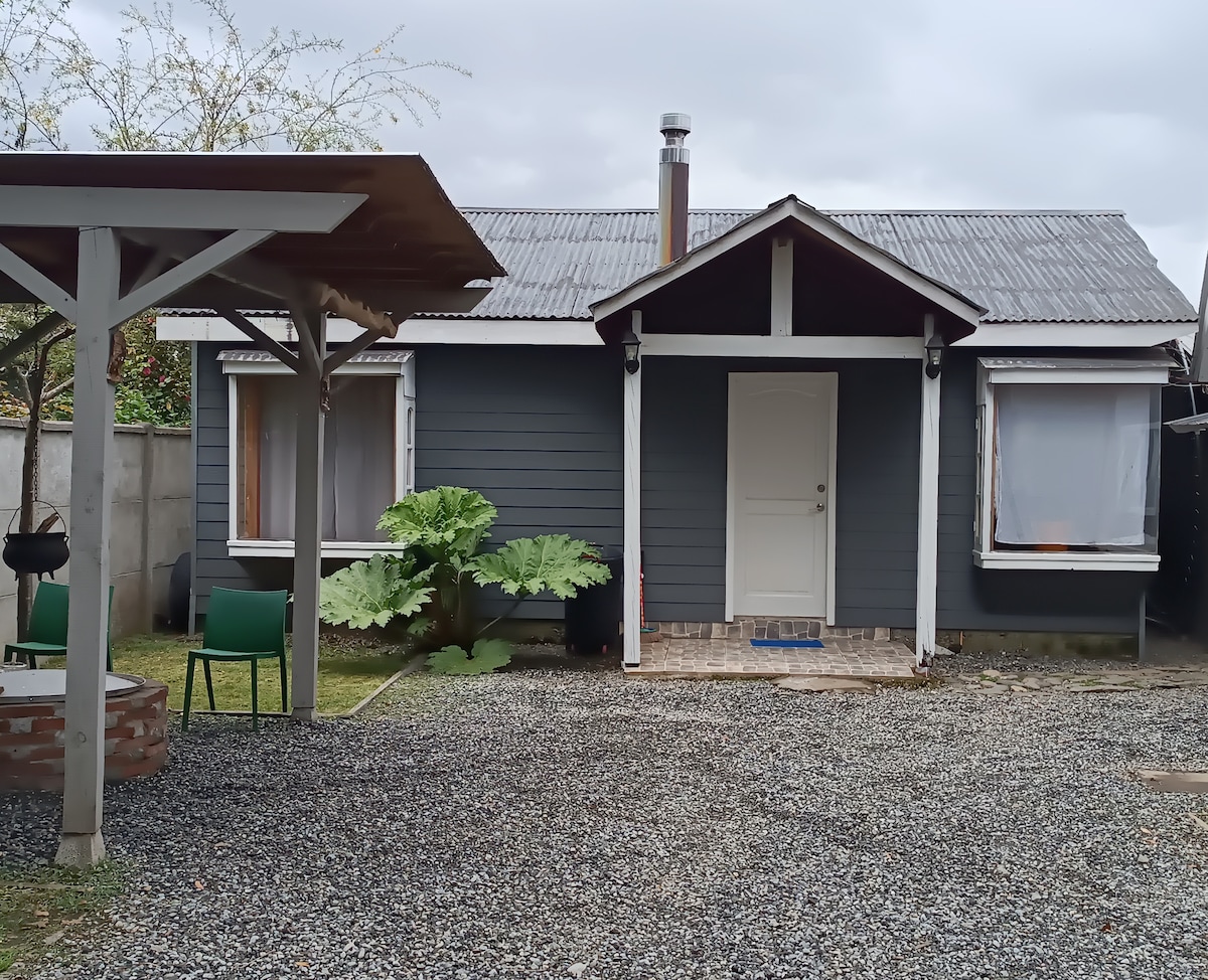 A charming cabin exterior is shown, with a wooden porch and a sloped roof. Large windows flank the entrance, revealing a welcoming atmosphere. Lush green foliage is positioned beside the front, and gravel pathways lead to the entrance.