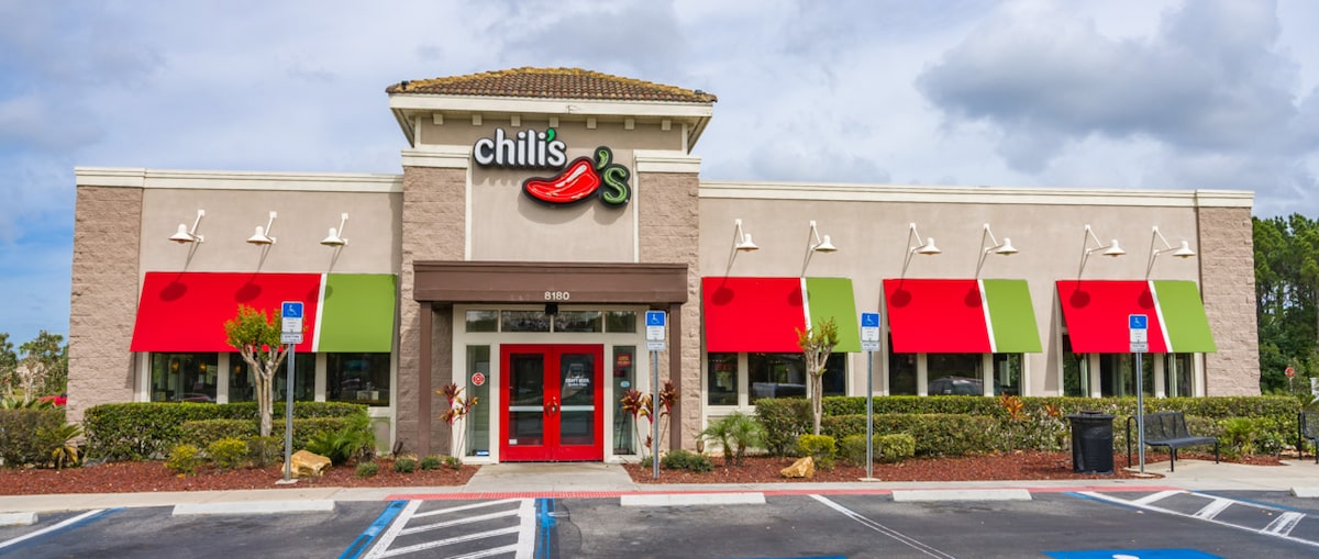 The image displays the exterior of a Chili's restaurant featuring a modern façade with a prominent red chili logo. Large windows are covered by bright red awnings, and landscape details including shrubs and potted plants enhance the entrance. Accessible parking spaces are visible in front.