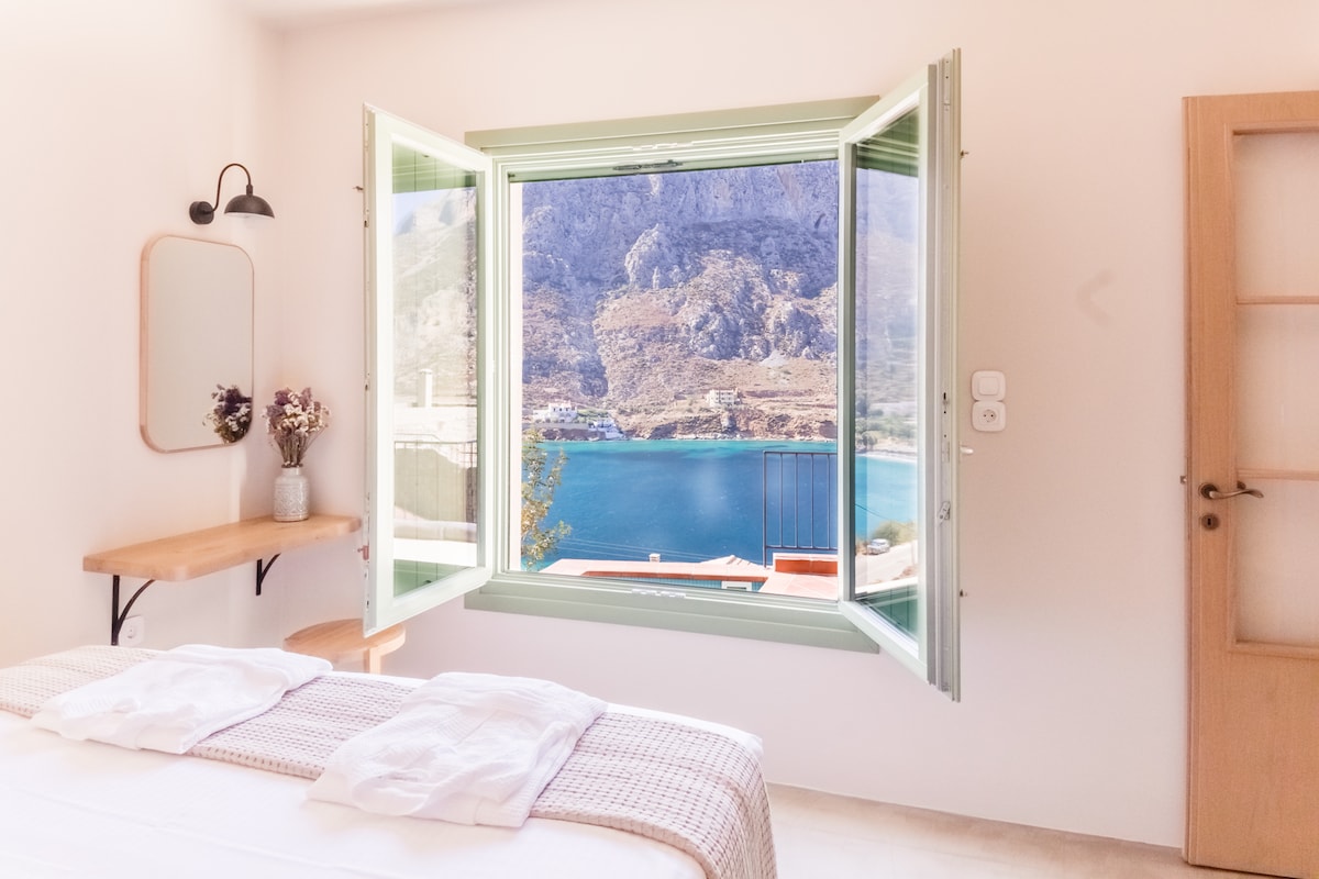 A bedroom features a spacious bed with crisp, white linens. A large window, framed in soft green, opens to reveal stunning views of the sea and surrounding mountains. A wooden shelf hangs nearby, adorned with a small vase of flowers, adding a touch of natural beauty.