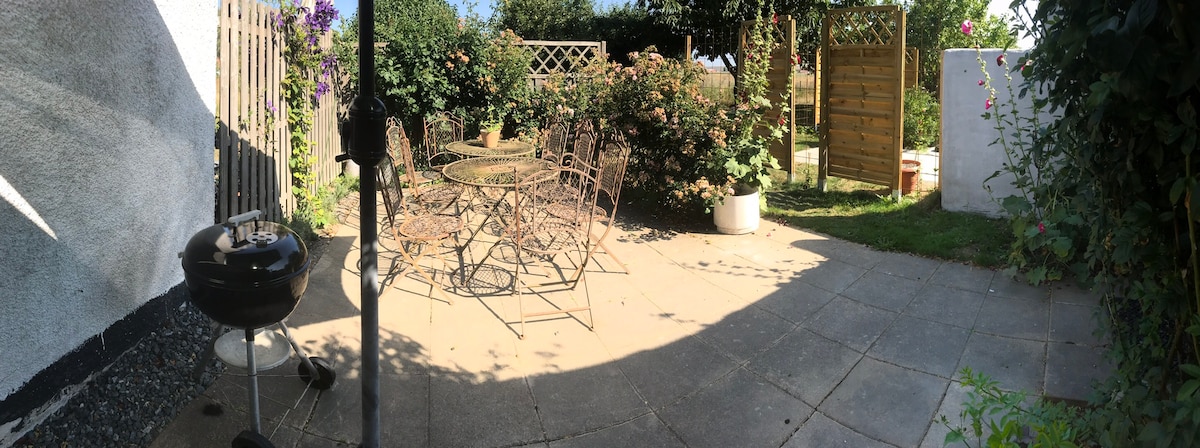 An outdoor patio features a stone floor surrounded by greenery, with a round dining table and several wrought iron chairs. A grill is positioned nearby, and climbing plants can be seen on a trellis, creating a private and inviting space for relaxation.