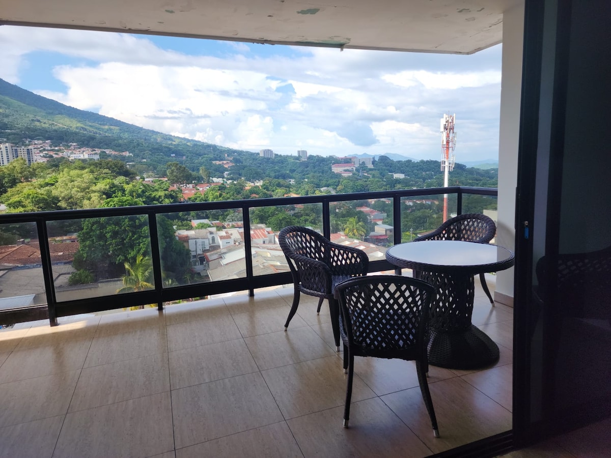 A spacious terrace features a round table surrounded by four woven chairs, offering views of the surrounding hills and cityscape. Large sliding doors provide access to the outdoor area, where bright skies and greenery can be observed, creating an inviting space for relaxation.