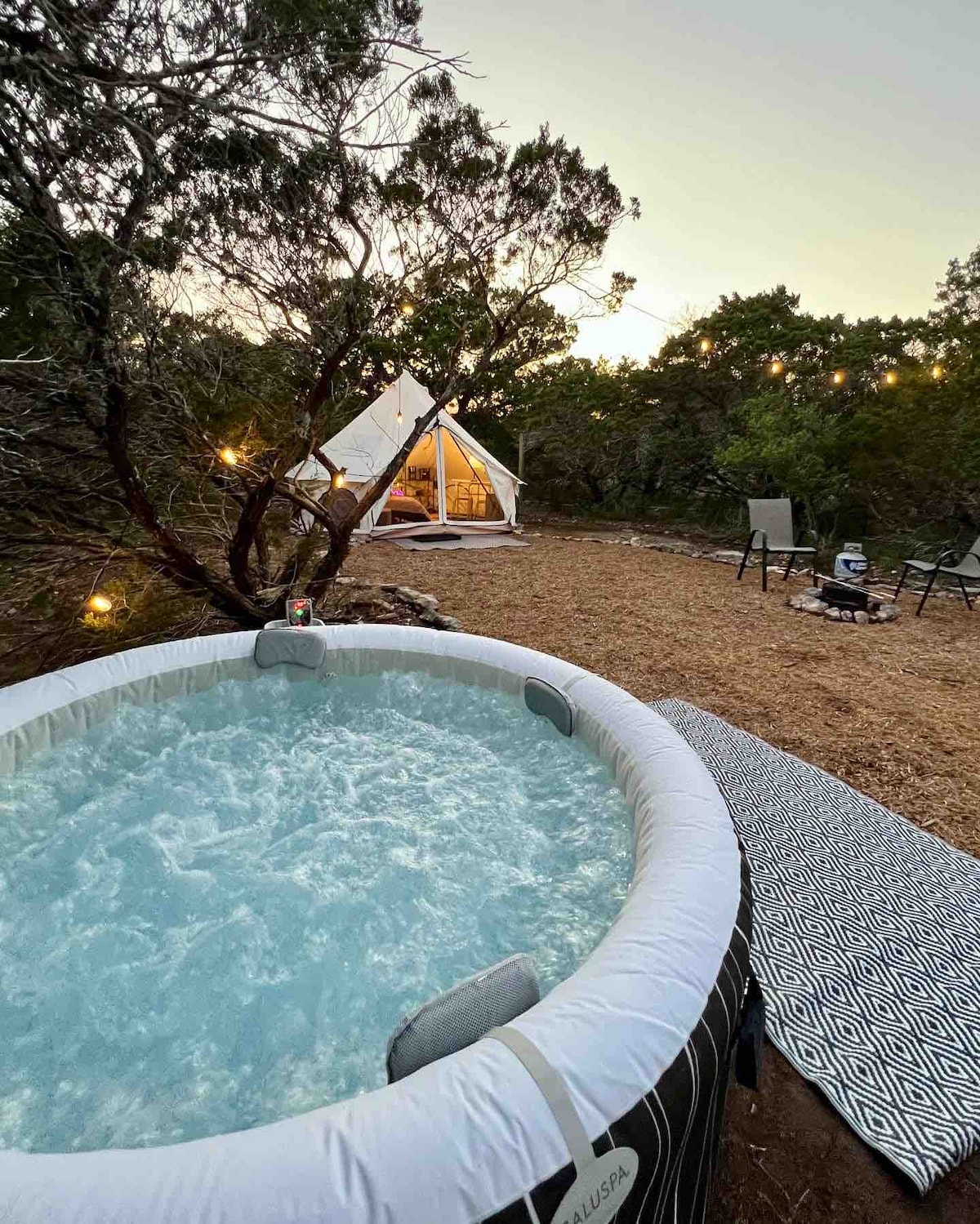 A secluded outdoor area features a private hot tub filled with bubbling water, situated adjacent to the tent. A patterned blanket lies on the ground, and two chairs are positioned nearby. Soft lighting from the surrounding trees enhances the tranquil setting.