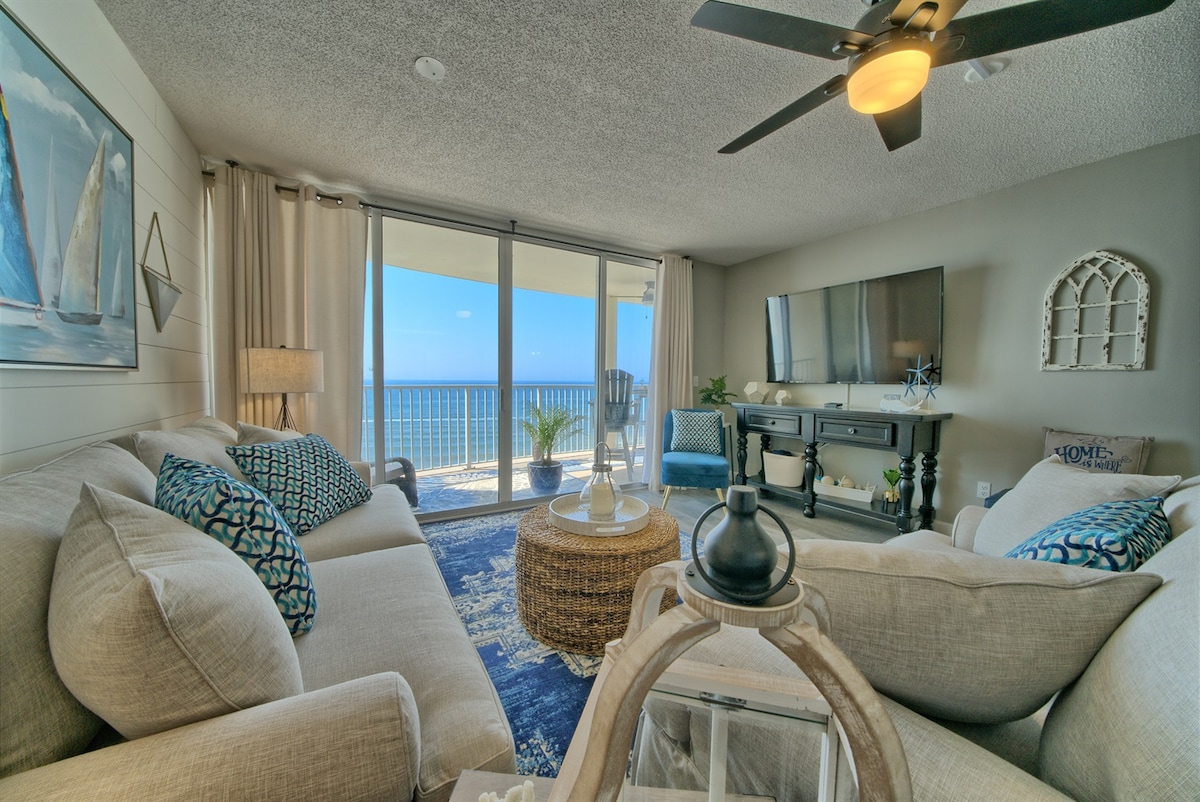 The living area features light-colored sofas adorned with decorative cushions, a round coffee table, and a flat-screen television. Large glass doors offer a clear view of the Gulf of Mexico and access to the spacious balcony, enhancing the room's open ambiance.