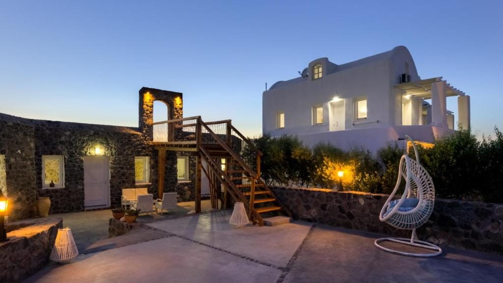 The exterior features a welcoming courtyard, with stone pathways leading to a two-story white building. A wooden staircase ascends from the terrace, surrounded by greenery. A hanging chair offers a relaxing space, illuminated by soft outdoor lighting at dusk.
