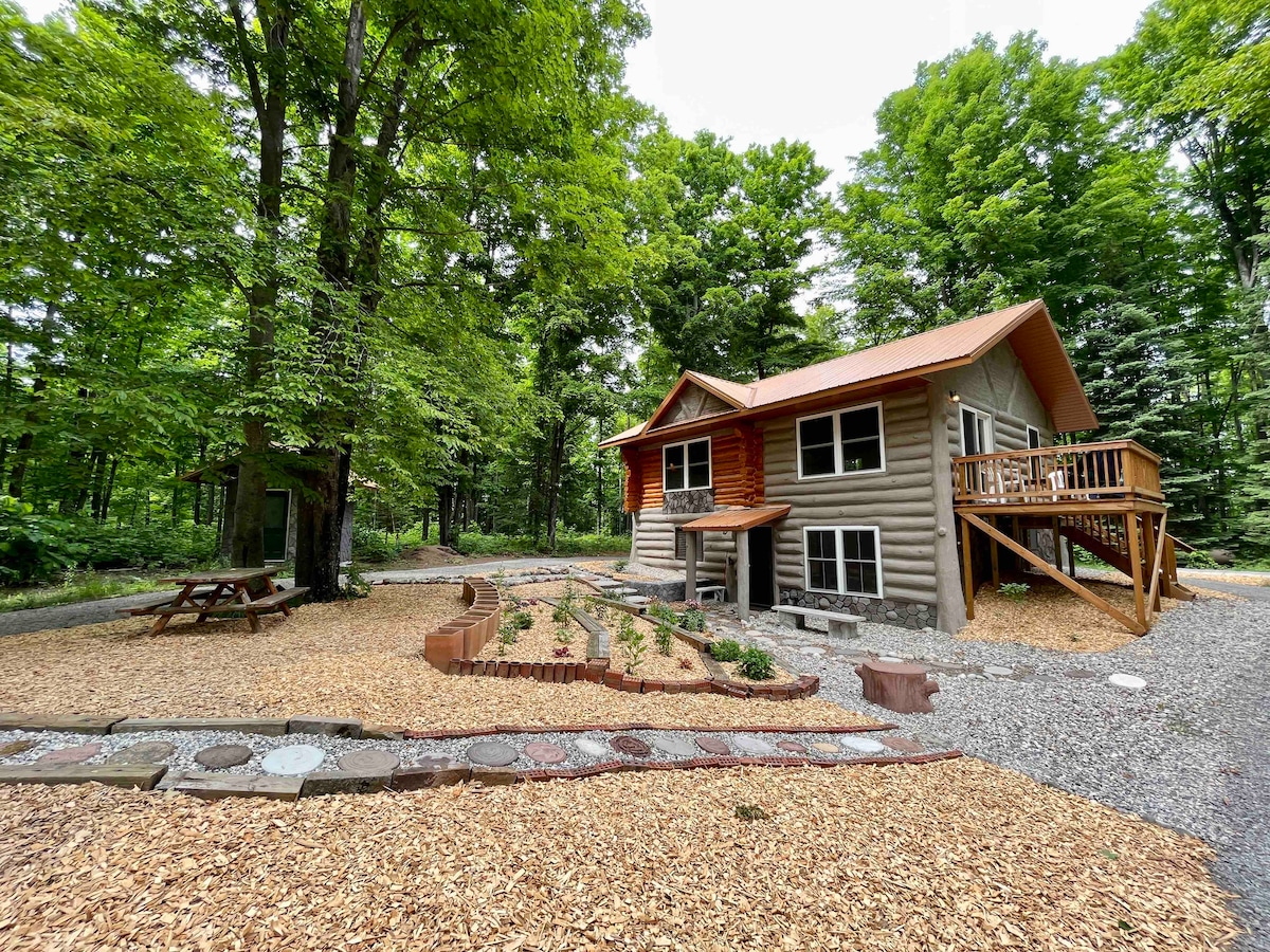 A log cabin is nestled amid lush greenery. The exterior features a combination of log walls and a sloped roof. A pathway made of stones leads to the entrance. Surrounding areas are landscaped with mulch and garden beds, while picnic tables invite outdoor relaxation.
