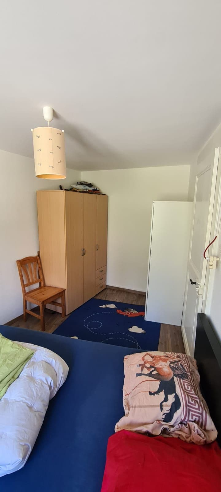 A simple bedroom displays a spacious bed with folded bedding and decorative pillows. Nearby, a wardrobe and a refrigerator are positioned against the wall. A wooden chair and a blue area rug featuring patterns are visible on the floor, completing the functional layout.