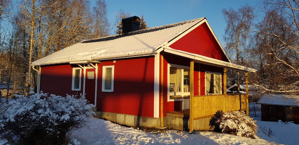 The exterior of the summer house is showcased, featuring a bright red facade with white trim under a clear blue sky. Snow blankets the ground, creating a stark contrast with the vibrant color. A wooden terrace extends from the front, complemented by surrounding snow-dusted shrubs.